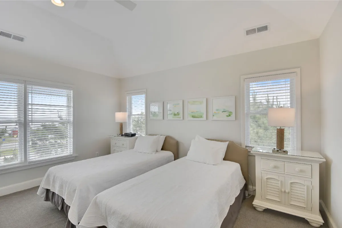 A bright, inviting bedroom with two twin beds positioned side by side, each topped with soft linens. Table lamps and framed artwork add charm, while a large window bathes the room in natural light. The arrangement is ideal for children or friends sharing the space.
