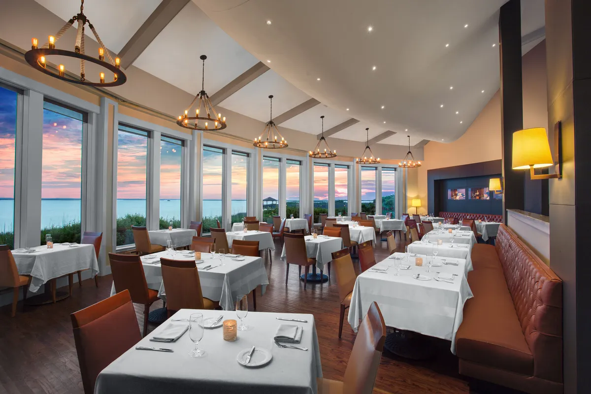 A spacious, elegant dining area with large windows overlooking a sunset view, chandeliers providing ambient lighting, dining tables set for guests, and a cozy sofa seating area. The setting offers a sense of comfort and luxury.