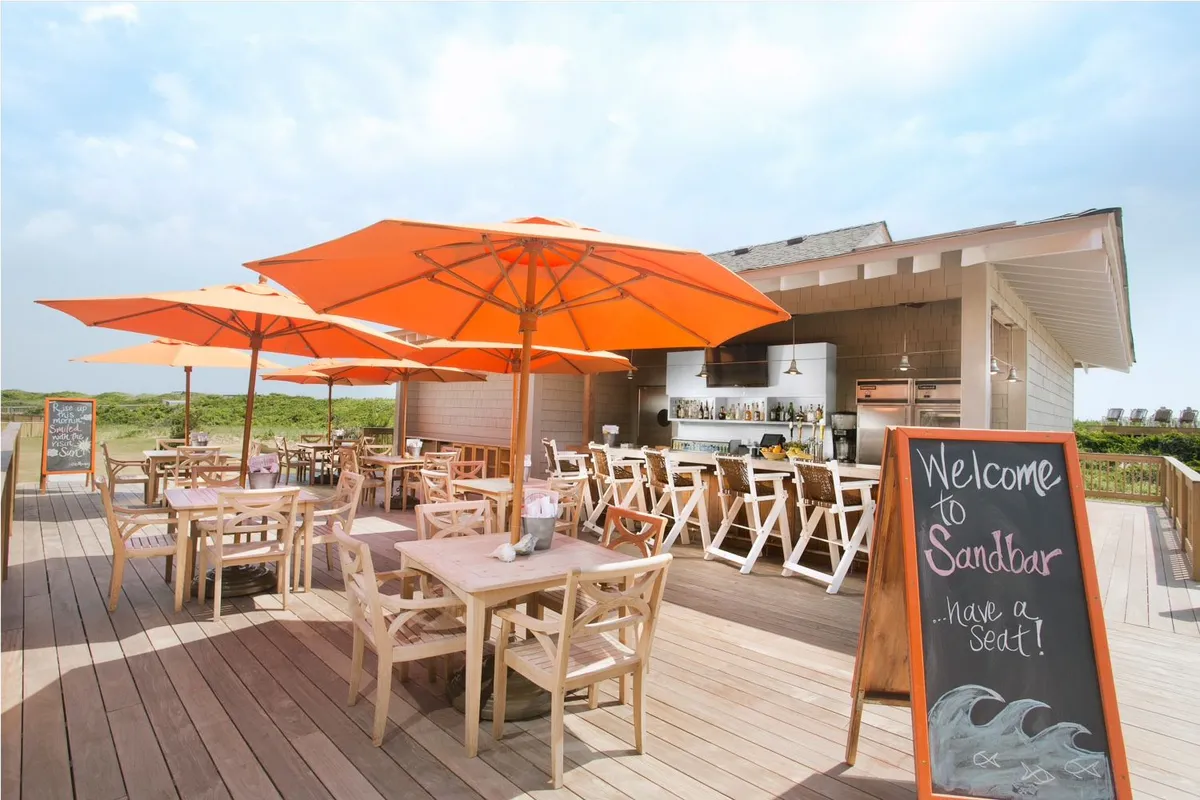 This image captures an inviting outdoor dining area with orange umbrellas, wooden tables, and a sandbar signage, suggesting a lively setting for group gatherings. People are seen enjoying meals and socializing, and the outdoor kitchen hints at the possibility of sunset happy hours and themed events.