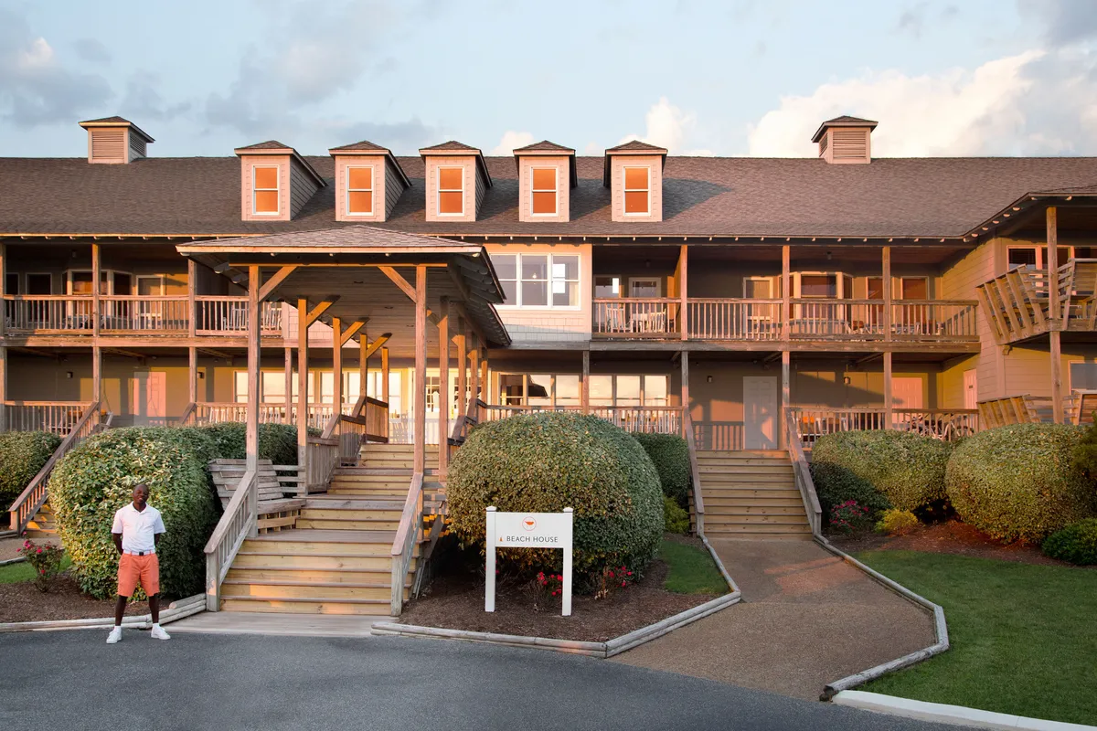 The main entrance to a beach house is shown, with a broad porch area framed by landscaped bushes and a beach house sign. The scene is bathed in warm, golden light, suggesting the ideal spot for morning coffee, evening gatherings, and tranquil moments with loved ones (including pets).