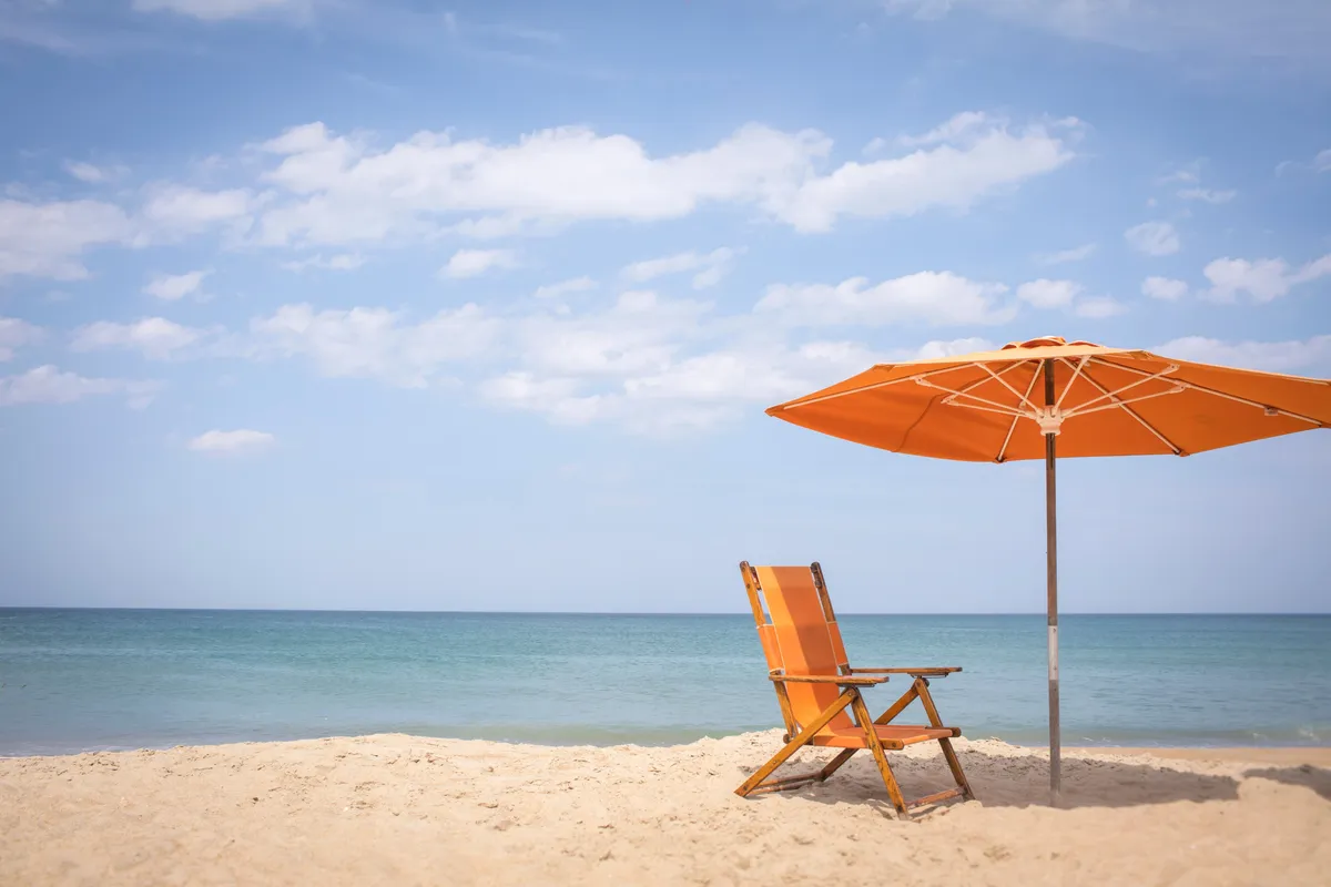 An idyllic beachfront scene with an orange beach chair and umbrella placed on soft sand overlooking a calm ocean. The setting is tranquil, and the scene invites relaxation, sunbathing, or family picnics just steps from the water.