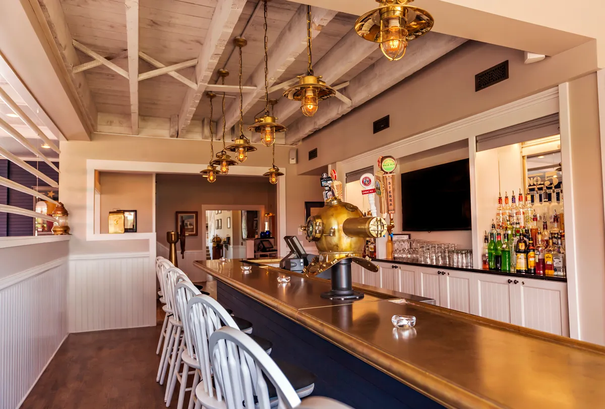 A stylish, lively bar with a gleaming brass bar top, modern hanging light fixtures, and rows of bar stools. Bottles of liquor are neatly arranged behind the bar, and guests are enjoying drinks, socializing, and watching sports on TV, creating a vibrant yet relaxed atmosphere perfect for unwinding with friends or celebrating special occasions.
