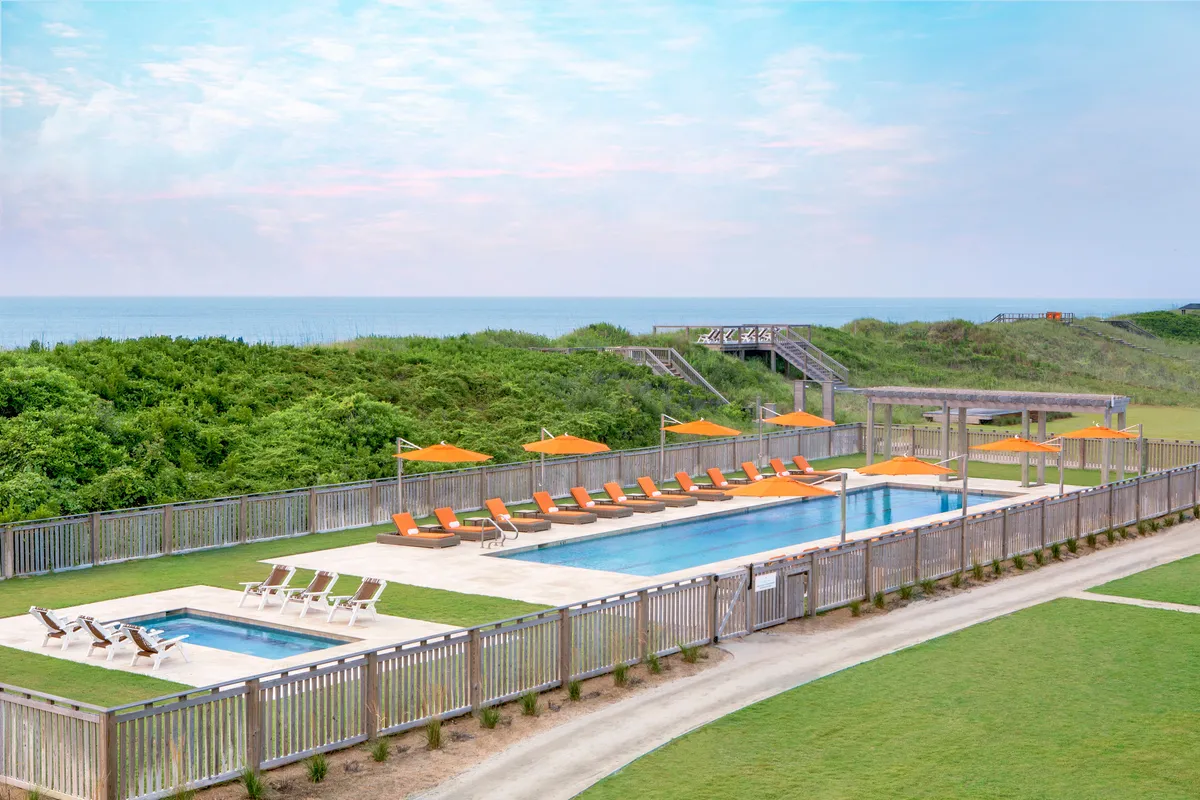 A serene exterior shot of a swimming pool surrounded by lounge chairs and orange umbrellas, with lush greenery and an expansive ocean view in the background. The balconies and porches of the property are visible, connecting the pool area to the private outdoor spaces above.