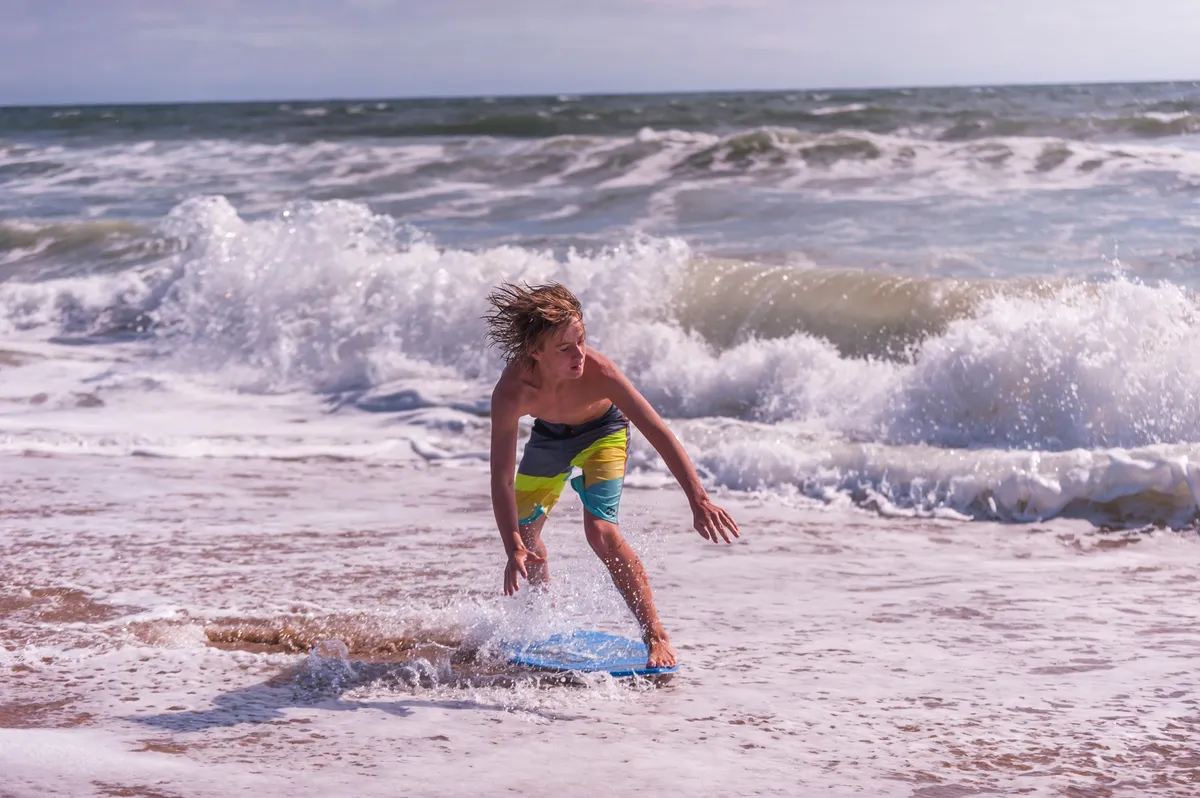 A joyful moment is captured as a boy rides a wave on a boogie board in the sunlit surf, with the expansive ocean stretching beyond. The energy is playful and spontaneous, highlighting active, memorable family adventures on the water.