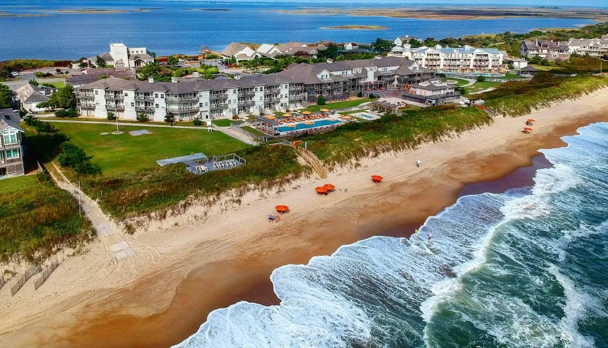 An inviting exterior scene of a beachfront property highlighting a sparkling swimming pool, orange beach umbrellas, and lush greenery adjacent to the ocean. Guests can be seen enjoying sunbathing and poolside relaxation with easy beach access.