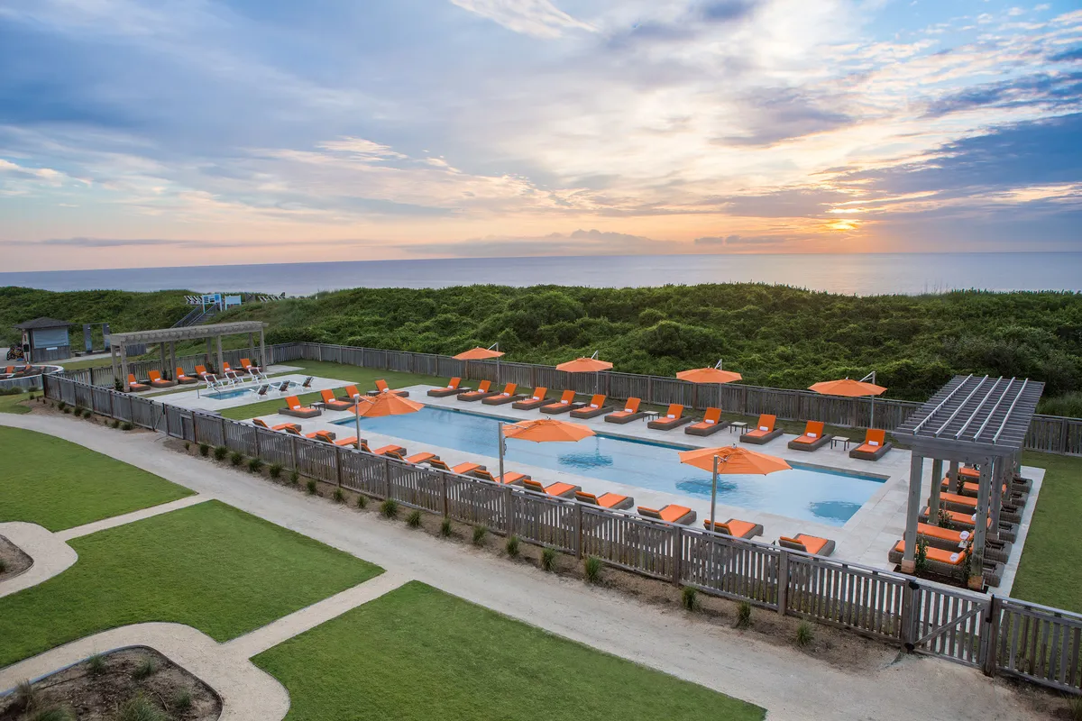 A spacious outdoor swimming pool lined with lounge chairs and orange umbrellas, with an ocean view and the sun setting in the background. The pool area exudes a serene and cheerful atmosphere, inviting for both relaxation and socializing.