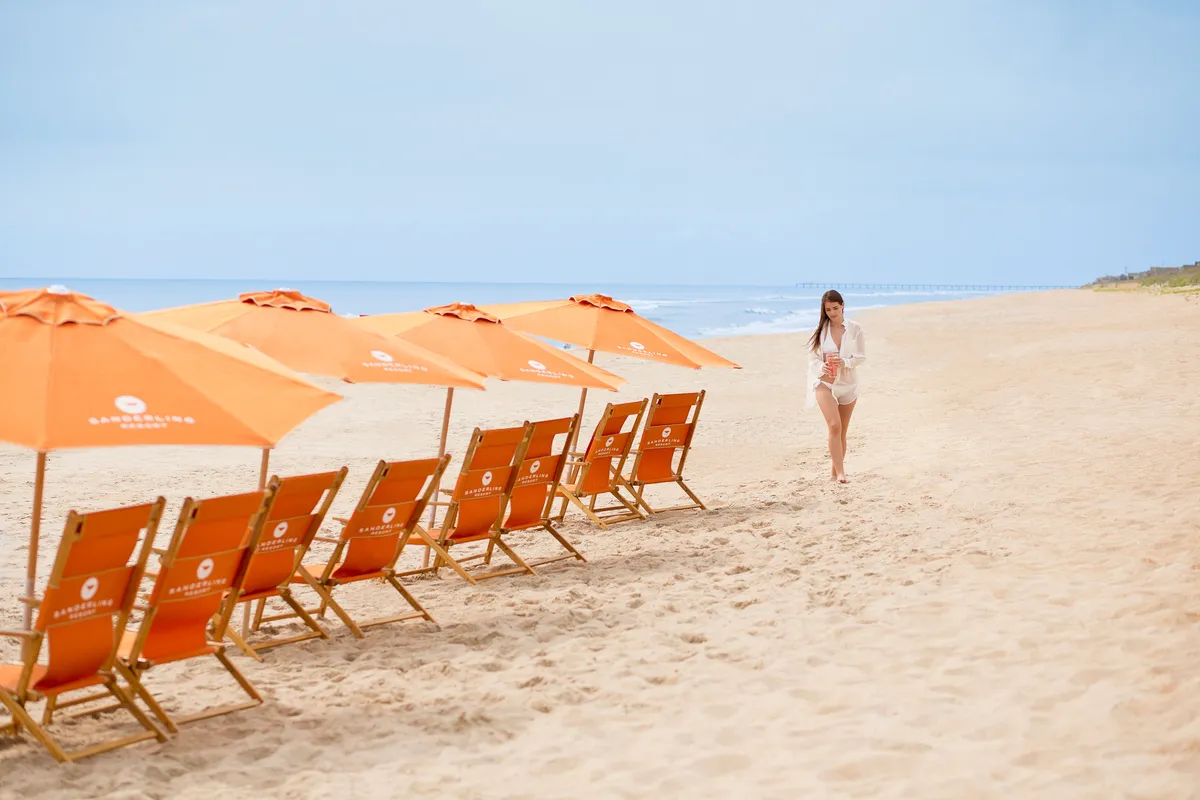 This image captures a serene beach scene with orange beach umbrellas and wooden beach chairs facing the ocean, while a person strolls along the shoreline. The sand looks soft and inviting, and the light suggests a warm, family-friendly day perfect for outdoor play and relaxation.