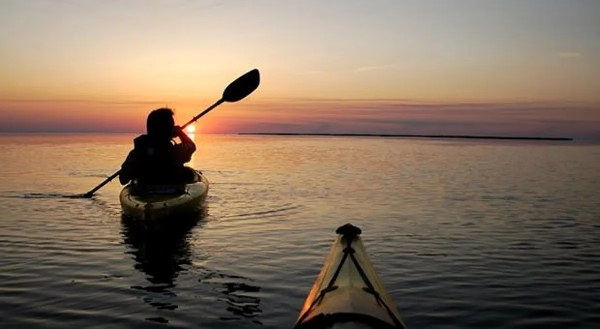 A tranquil scene featuring a kayaker paddling on calm waters during sunset, with the sky painted in warm hues and the water surface reflecting the colors. The overall mood captures a connection to nature and the outdoors.