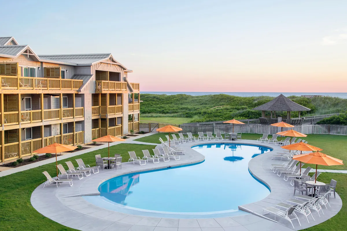 The image features a resort pool area with inviting blue water, orange umbrellas, and balconies overlooking the scene. Sunbathers and families might gather here to swim or relax after a day at the beach, with a calm, inviting ambiance that echoes resort luxury.