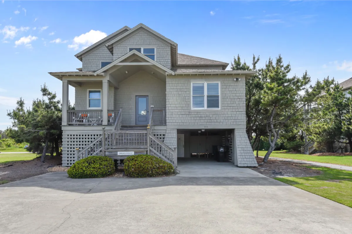The image showcases the inviting facade of a coastal single-family house in Duck, North Carolina. The front porch is accented with cozy seating, perfect for socializing or soaking in vibrant sunsets, while the landscaped yard adds to the home's charm and curb appeal. The scene evokes a sense of warmth and relaxation, with ample space for family gatherings and moments of outdoor serenity.